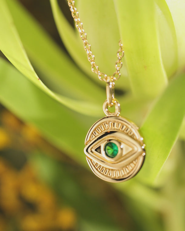 Closeup video of an ENVYONE 18K Gold Plated evil eye pendant necklace with a dark green cubic zirconia stone against lush green leaves.