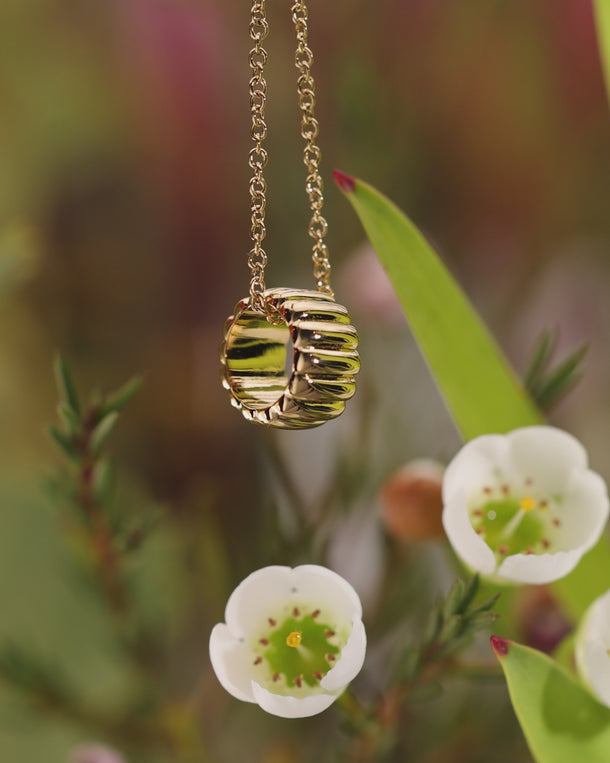 Closeup video of ENVYONE 18K Gold-Plated necklace featuring a sculptural ribbed pendant displayed among flowers.