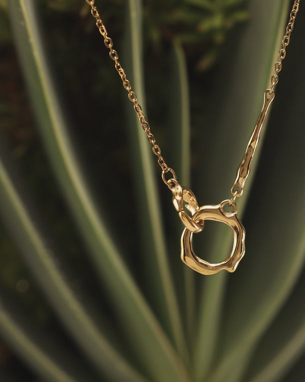 Closeup of an ENVYONE 18K Gold Plated necklace with sculptural interlocking links styled against dark green foliage.